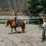 Woman training rider on horse in a sunny outdoor arena with natural surroundings.